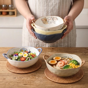 Peut inclure: Une personne tient une pile de trois bols en c&eacute;ramique cr&egrave;me, jaune et bleu. Le bol sup&eacute;rieur pr&eacute;sente un motif de chat. Deux bols sont remplis de nourriture, l'un de salade et l'autre de nouilles, de crevettes et de l&eacute;gumes. Les bols sont sur des nattes tress&eacute;es.