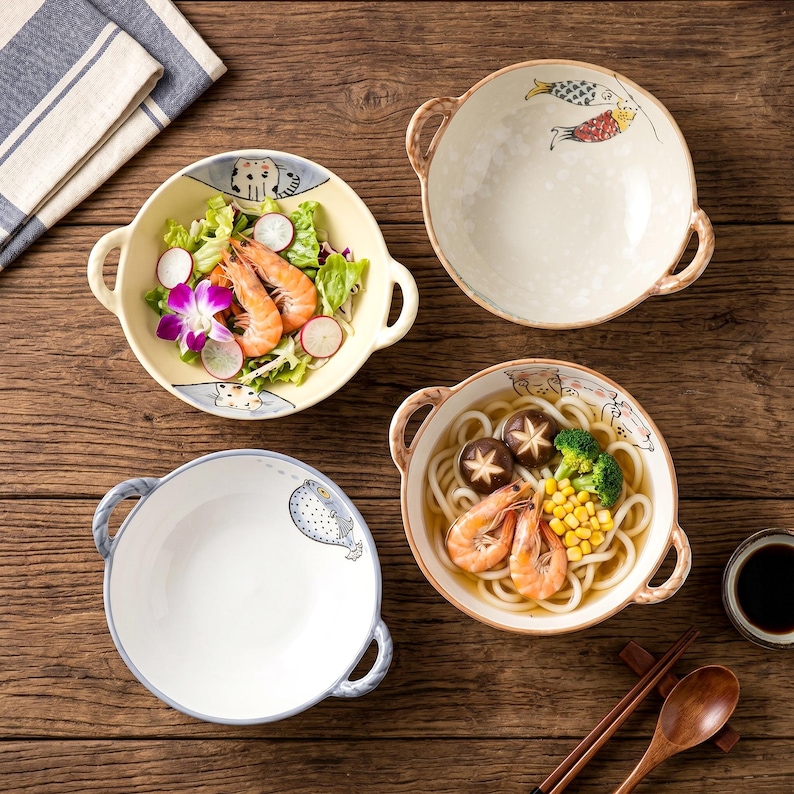Peut inclure: Quatre bols en c&eacute;ramique avec des poign&eacute;es, chacun rempli de nourriture. L'un contient une salade avec des crevettes, un autre des nouilles udon avec des crevettes, et les deux autres sont vides. Les bols ont des motifs d&eacute;coratifs de poissons et de chats.