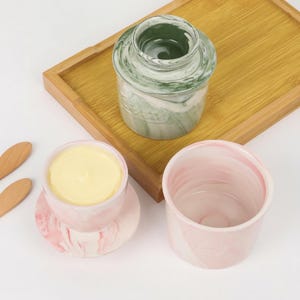 Puede incluir: Una colecci&oacute;n de mantequilleras y recipientes de cer&aacute;mica. Una mantequillera veteada verde y blanca con tapa, una mantequillera veteada rosa y blanca con mantequilla dentro y un recipiente vac&iacute;o a juego. Los art&iacute;culos est&aacute;n sobre una bandeja de madera.