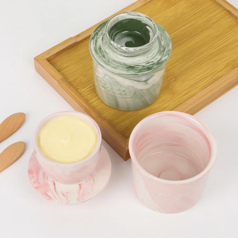Puede incluir: Una colecci&oacute;n de mantequilleras y recipientes de cer&aacute;mica. Una mantequillera veteada verde y blanca con tapa, una mantequillera veteada rosa y blanca con mantequilla dentro y un recipiente vac&iacute;o a juego. Los art&iacute;culos est&aacute;n sobre una bandeja de madera.