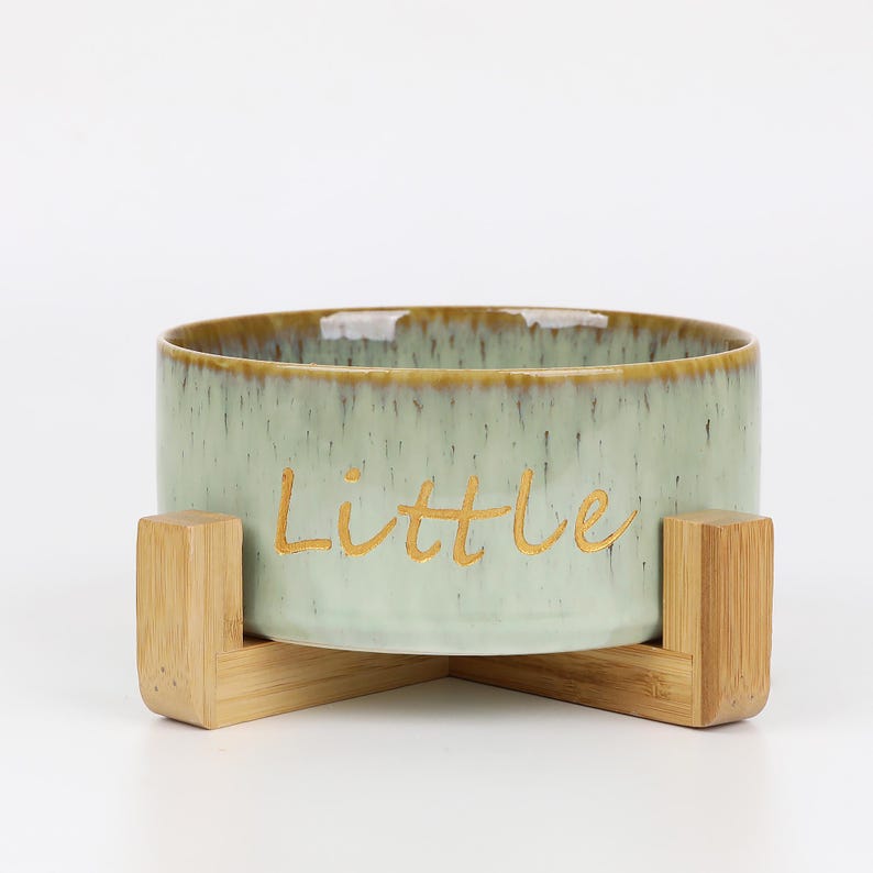 May include: A light green ceramic pet bowl with the word "Little" in gold lettering. The bowl sits on a natural wood stand. The bowl has a light brown rim.