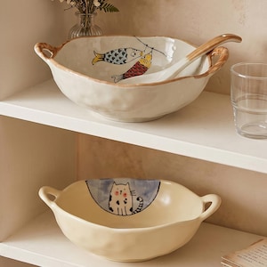Peut inclure: Deux bols en c&eacute;ramique avec des poign&eacute;es sur une &eacute;tag&egrave;re blanche. Le bol sup&eacute;rieur pr&eacute;sente un motif de poisson et une cuill&egrave;re est &agrave; l'int&eacute;rieur. Le bol inf&eacute;rieur pr&eacute;sente une illustration de chat. Les deux bols sont de couleur cr&egrave;me et sont adapt&eacute;s au service.