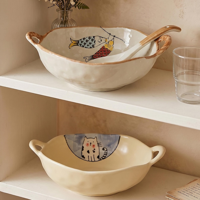 Peut inclure: Deux bols en c&eacute;ramique avec des poign&eacute;es sur une &eacute;tag&egrave;re blanche. Le bol sup&eacute;rieur pr&eacute;sente un motif de poisson et une cuill&egrave;re est &agrave; l'int&eacute;rieur. Le bol inf&eacute;rieur pr&eacute;sente une illustration de chat. Les deux bols sont de couleur cr&egrave;me et sont adapt&eacute;s au service.