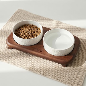 Peut inclure: Un support de gamelle pour animaux en bois avec deux bols en céramique blancs. Un bol est rempli de nourriture sèche pour animaux, l'autre d'eau. Les bols ont un motif floral. Le support est gravé du mot "Klee" et d'une empreinte de patte.