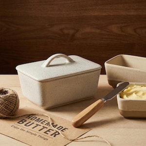 May include: A speckled, off-white ceramic butter dish with a lid and handle, alongside a butter knife with a wooden handle. A small dish holds butter. A tag reads "Homemade Butter" and "Enjoy the Simple Things."