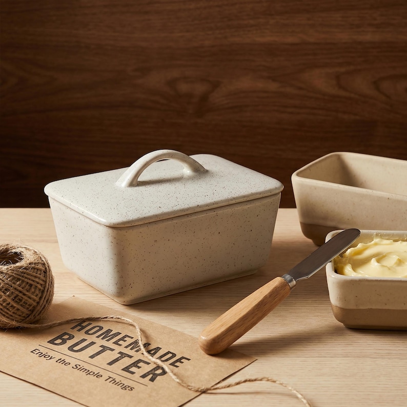 May include: A speckled, off-white ceramic butter dish with a lid and handle, alongside a butter knife with a wooden handle. A small dish holds butter. A tag reads "Homemade Butter" and "Enjoy the Simple Things."