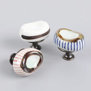 May include: Three decorative ceramic cabinet knobs. One knob is white with a brown stripe, another is white with blue stripes, and the third has red stripes. Each knob is mounted on a dark metal base.