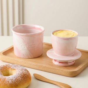 Puede incluir: Mantequera de cer&aacute;mica rosa y blanca veteada con tapa, llena de mantequilla amarilla, y un recipiente abierto a juego. El juego est&aacute; sobre una bandeja de madera, junto a un bagel y un cuchillo de mantequilla de madera.