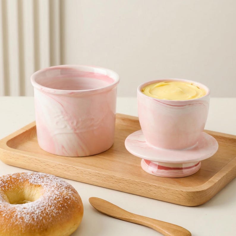 Puede incluir: Mantequera de cer&aacute;mica rosa y blanca veteada con tapa, llena de mantequilla amarilla, y un recipiente abierto a juego. El juego est&aacute; sobre una bandeja de madera, junto a un bagel y un cuchillo de mantequilla de madera.