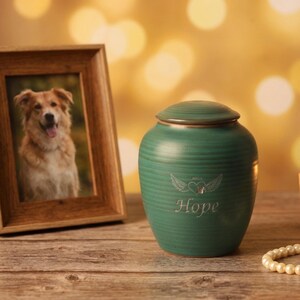 May include: A green ceramic pet urn with a lid, engraved with wings, a heart, and the word "Hope." A framed photo of a dog is to the left. The urn is on a wooden surface.