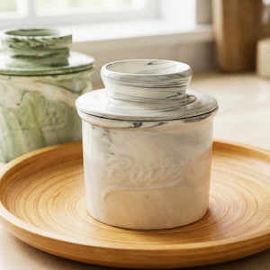 Puede incluir: Una mantequera blanca y gris con dise&ntilde;o de m&aacute;rmol y tapa, sobre una bandeja de madera. La palabra "Butter" est&aacute; grabada en relieve en la parte delantera. Otro plato verde con dise&ntilde;o de m&aacute;rmol en el fondo.
