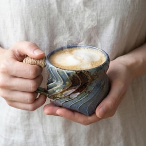 Pode incluir: Uma caneca de cerâmica artesanal com esmalte azul e castanho, cheia de uma bebida de café espumosa. A caneca tem um design único e texturizado e uma alça tecida. O vapor sobe do café, sugerindo que está quente.