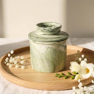 Puede incluir: Una mantequera de cer&aacute;mica veteada verde y blanca con tapa, colocada sobre una bandeja redonda de madera. El recipiente tiene una base cil&iacute;ndrica y una tapa a juego. Flores blancas y vegetaci&oacute;n dispuestas alrededor.