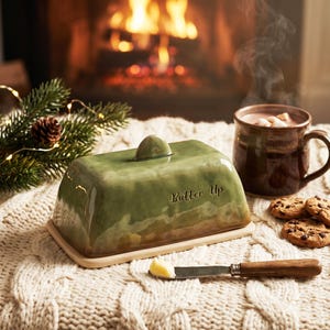 Op de afbeelding: Een groene keramische boterdoos met de tekst "Butter Up" op een crèmekleurige basis. Een klein klontje boter ligt op een mes met een houten handvat. Een mok warme chocolademelk en koekjes zijn in de buurt.