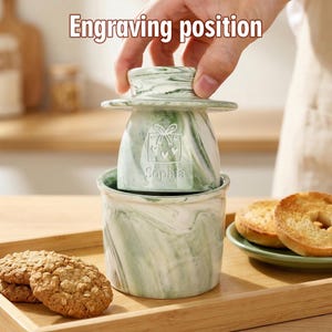 Puede incluir: Una mantequera con dise&ntilde;o de m&aacute;rmol verde y blanco con tapa, con un dise&ntilde;o de caja de regalo y el nombre "Sophia". El plato tiene una base a juego. El texto "Engraving position" est&aacute; en la parte superior. Galletas y bagels en el fondo.