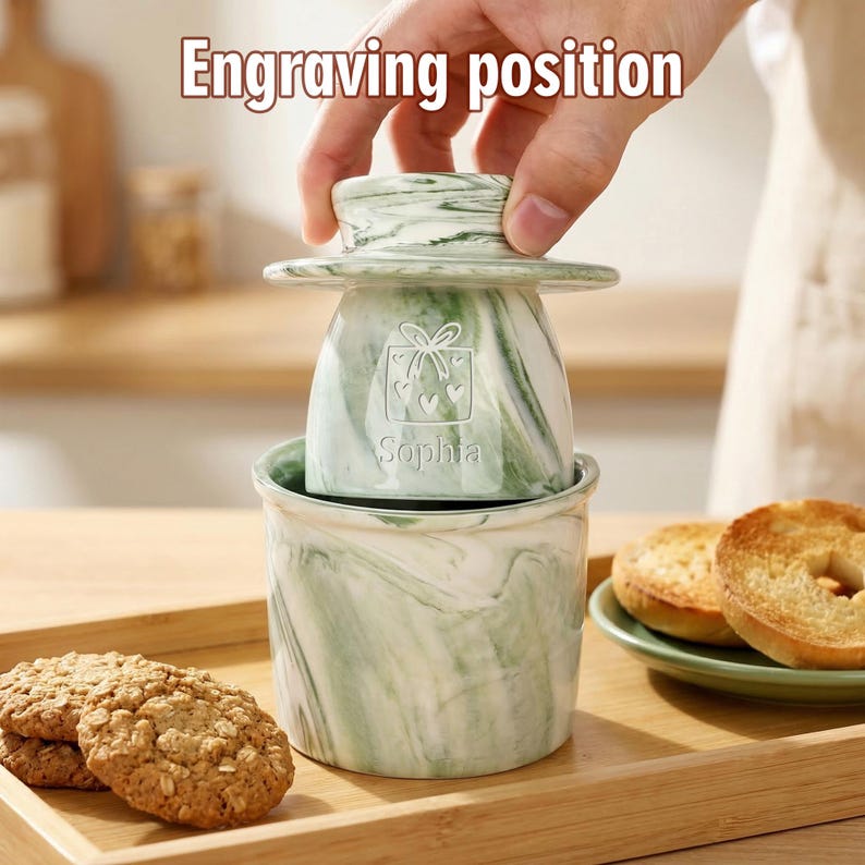 Puede incluir: Una mantequera con dise&ntilde;o de m&aacute;rmol verde y blanco con tapa, con un dise&ntilde;o de caja de regalo y el nombre "Sophia". El plato tiene una base a juego. El texto "Engraving position" est&aacute; en la parte superior. Galletas y bagels en el fondo.