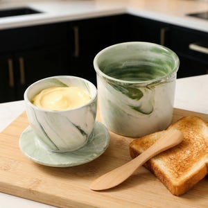 Puede incluir: Un juego de mantequillera con un recipiente a juego, un platillo y un cuchillo de mantequilla de madera. La mantequillera y el recipiente tienen un dise&ntilde;o de m&aacute;rmol verde y blanco. Dos rebanadas de pan tostado est&aacute;n al lado del cuchillo sobre una tabla de cortar de madera.