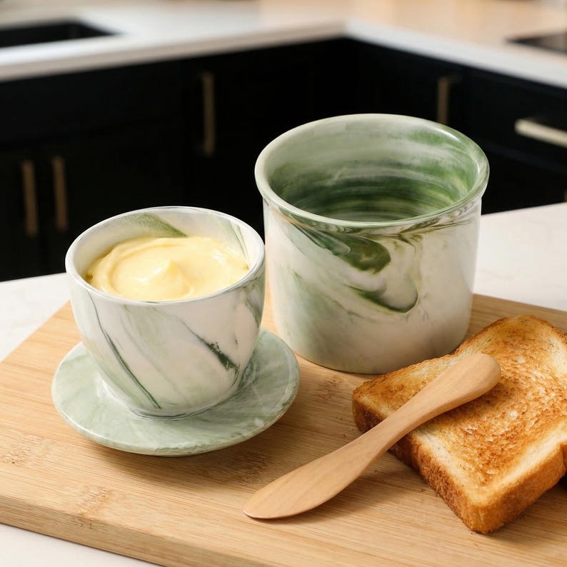 Puede incluir: Un juego de mantequillera con un recipiente a juego, un platillo y un cuchillo de mantequilla de madera. La mantequillera y el recipiente tienen un dise&ntilde;o de m&aacute;rmol verde y blanco. Dos rebanadas de pan tostado est&aacute;n al lado del cuchillo sobre una tabla de cortar de madera.