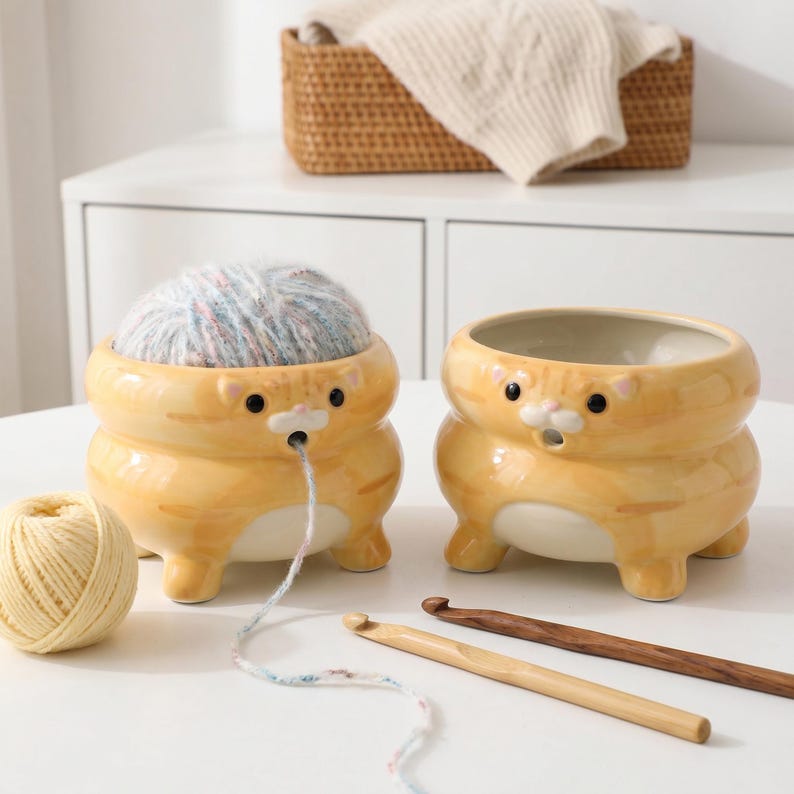 以下が含まれることがあります： オレンジ色の猫の形をした陶器製の毛糸入れが2つ。1つは毛糸が入っており、もう1つは空です。黄色の毛糸玉と2本の木製の編み棒も写っています。