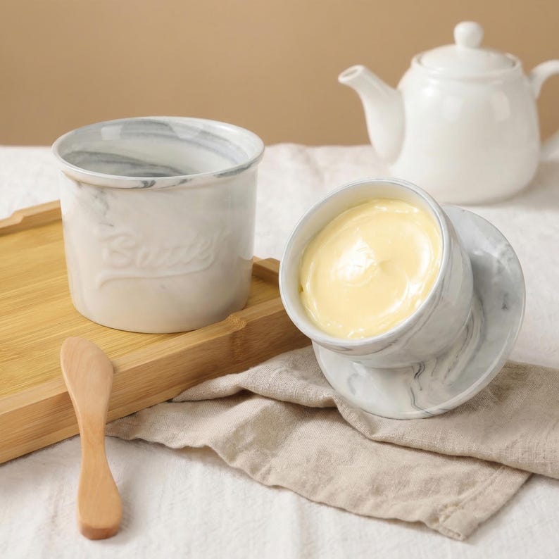 Puede incluir: Un mantequero blanco con dise&ntilde;o de m&aacute;rmol y tapa, lleno de mantequilla amarilla. Un recipiente a juego con la palabra "Butter" en relieve en el lateral se encuentra en una bandeja de madera, junto con un cuchillo de mantequilla de madera. Una tetera blanca est&aacute; en el fondo.