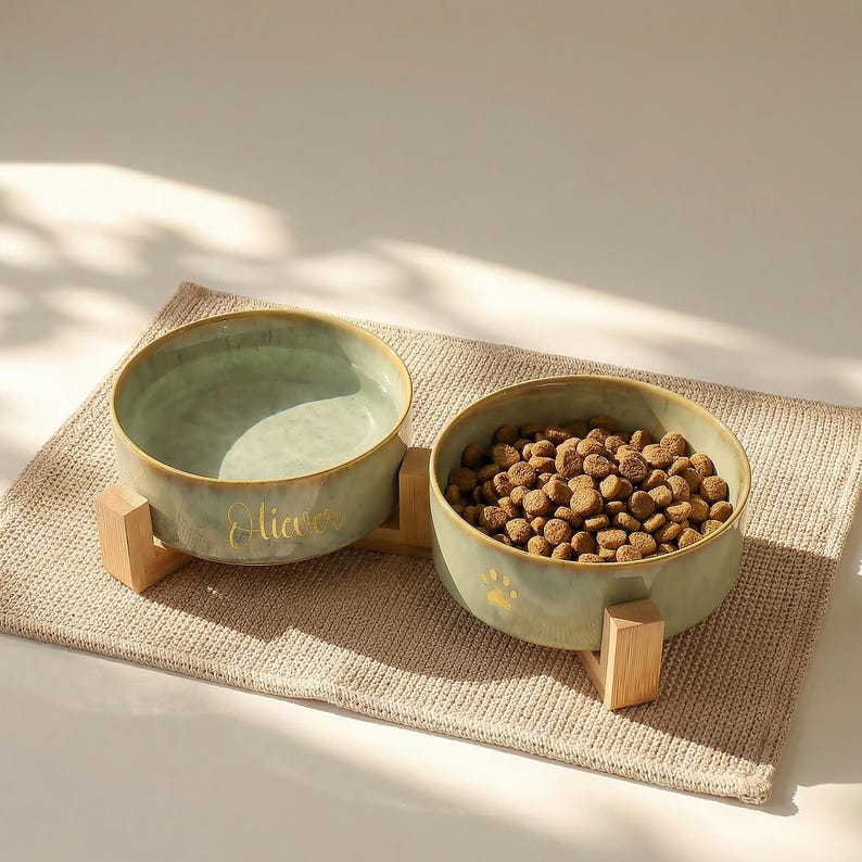 May include: A set of two ceramic pet bowls in a muted green hue, each resting on a wooden stand. One bowl is filled with dry pet food, the other is empty. The word "Oliver" is written on one bowl in gold lettering, and a paw print is on the other.