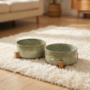May include: Two sage green ceramic pet bowls with wooden stands. One bowl has the name "Daisy" in gold lettering, and the other has a gold paw print. The bowls are on a fluffy white rug.