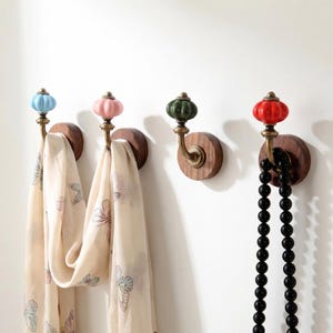 Puede incluir: Cuatro perchas de pared decorativas con pomos de cerámica de colores azul, rosa, verde y rojo. Cada gancho tiene un brazo de latón y una base de madera redonda. Dos ganchos sostienen bufandas estampadas y uno sostiene un collar de cuentas negro.