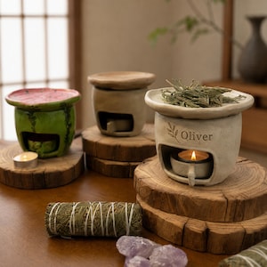Puede incluir: Tres quemadores de aceite de cerámica con velas de té en su interior. Uno es verde con la parte superior rosa, dos son beige. Un quemador beige presenta el nombre Oliver y un icono de hoja. Un manojo de hierbas secas y cristales morados.