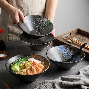 以下が含まれることがあります： 黒いセラミックラーメン丼のセット。1つの丼にはラーメン、エビ、野菜が入っています。別の丼は青と白の斑点模様です。日本のスタイルの食器セットの一部です。