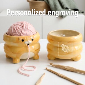 以下が含まれることがあります： ピンクの糸が入った猫のデザインのセラミック糸入れが2つ。ボウルはオレンジ色で、黒い目と白いお腹があります。2つ目のボウルには糸と針のグラフィックがあります。木製の編み棒も表示されています。