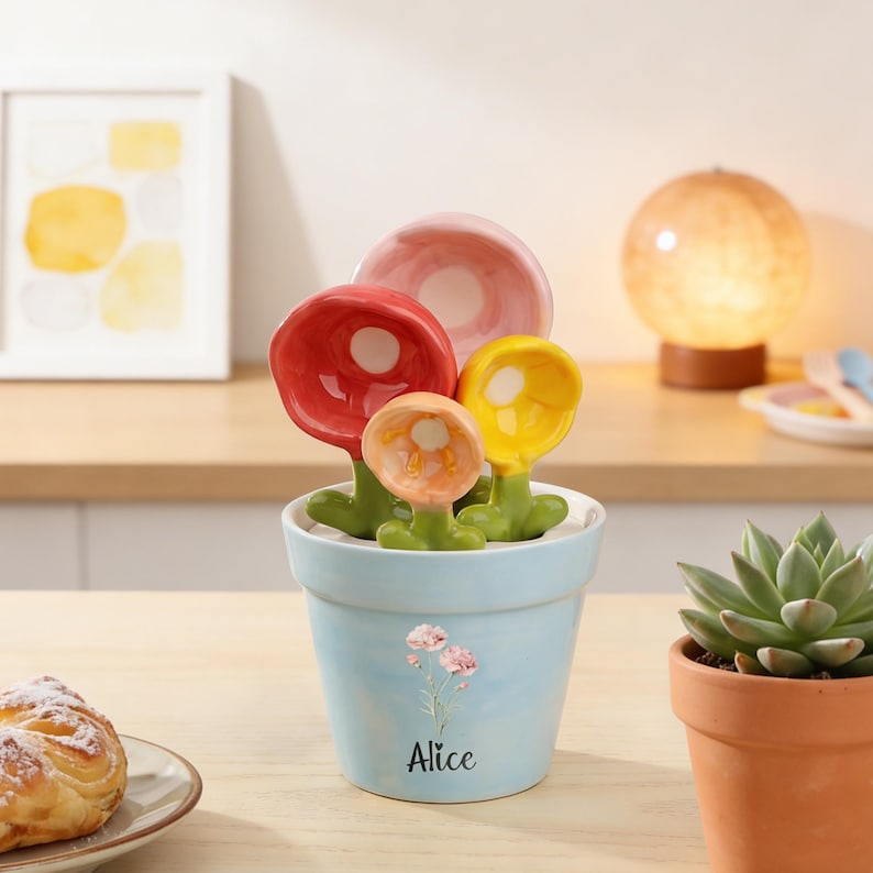 May include: A light blue ceramic flower pot with a floral design and the name "Alice" printed on the front. The pot holds four colorful ceramic flower measuring spoons in red, pink, yellow, and orange. A succulent plant in a terracotta pot is to the right.