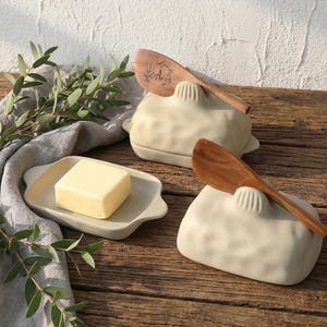 Op de afbeelding: Een boterservies met een lichtbeige keramische boterhouder en een houten botermes. Een blok boter ligt op een bijpassende serveerschaal. De set staat op een houten oppervlak met groen.