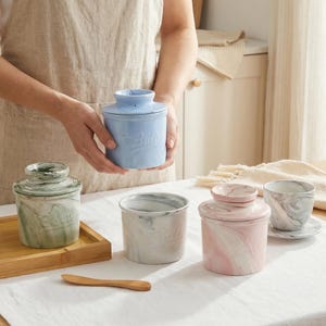 Puede incluir: Una colecci&oacute;n de mantequilleras de cer&aacute;mica en varios colores, incluyendo azul, verde, rosa y gris. Cada mantequillera tiene un dise&ntilde;o veteado y una tapa. Una mantequillera azul es sostenida por una persona. Tambi&eacute;n hay un cuchillo de mantequilla de madera.