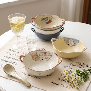 Peut inclure: Plusieurs bols en c&eacute;ramique avec des poign&eacute;es, d&eacute;cor&eacute;s de motifs peints &agrave; la main de poissons et de chats. Les bols sont de couleurs cr&egrave;me, bleu et jaune. Une cuill&egrave;re en c&eacute;ramique et un verre de vin sont &eacute;galement pr&eacute;sents.