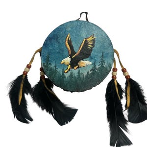 May include: A circular hand drum featuring a painted scene of a bald eagle in flight over a dark green forest. The drum is decorated with four leather strips adorned with small wooden beads and long black feathers hanging from the sides.