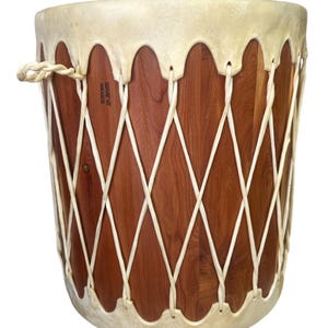 May include: A cylindrical drum with a reddish-brown wood grain body and a cream-colored rawhide head. The drum features a crisscross lacing pattern made of white cordage that secures the head to the base, with a small loop handle on the side.