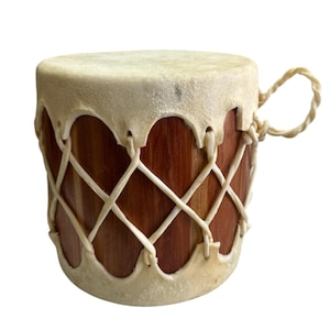 May include: A cylindrical drum featuring a reddish-brown wooden body wrapped in a white rawhide lacing pattern. The top and bottom edges are finished with thick, cream-colored rawhide, and a small braided rawhide loop extends from the side.