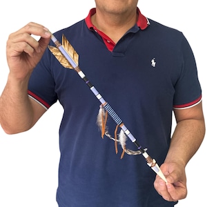 May include: A decorative arrow featuring a shaft with alternating light blue, black, and white bands. The arrow includes a fletching made of golden-brown feathers, a section wrapped in blue thread, and two dangling brown leather cords with white feathers.