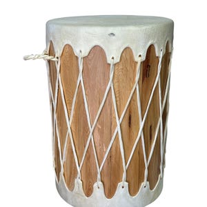 May include: A cylindrical wooden drum featuring a light brown grain finish. The drum is secured with white rope laced in a crisscross pattern between two white leather heads at the top and bottom. A small white rope handle is attached to the side.