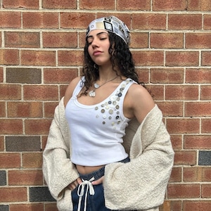 Puede incluir: Un top sin mangas blanco adornado con varios botones, combinado con una rebeca color crema y vaqueros azul oscuro. Un pañuelo estampado y un collar completan el look. La modelo está de pie frente a una pared de ladrillos.