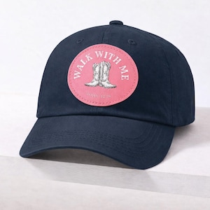 Puede incluir: Gorra de béisbol azul marino con un parche circular rosa. El parche presenta un dibujo de botas de vaquero y el texto "WALK WITH ME" y "JOHN 12:16". Una gorra clásica e informal para el uso diario.