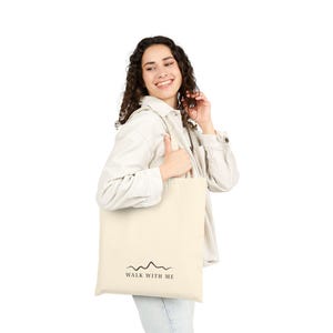 May include: Cream-colored tote bag with the text "WALK WITH ME" and a mountain range graphic in black. The bag is held by a person wearing a white button-down shirt and blue jeans.