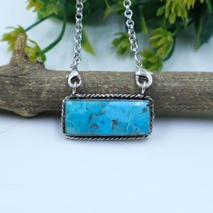 May include: A rectangular turquoise pendant necklace with a silver-colored chain. The turquoise stone is a vibrant blue with brown veining, set in a silver frame with a twisted rope design. The necklace is displayed against a natural wood background.