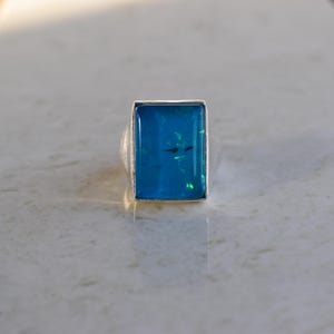 May include: A silver ring featuring a rectangular, vibrant blue stone set in a smooth metal bezel. The stone displays internal flecks of green and light blue, creating a luminous effect within the deep blue surface of the rectangular gemstone.
