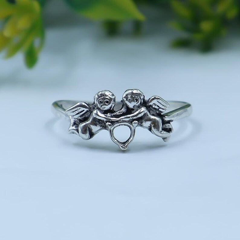 May include: A silver-toned ring featuring two cherub figures facing each other with wings extended. The cherubs are connected at the center by a small, open heart-shaped detail. The metal has a polished finish with darkened recessed areas.