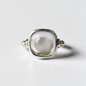 May include: A silver ring with a large, square, clear stone. The ring has gold accents on the sides. The stone is set in a silver bezel. The ring is on a white background. Jewelry.