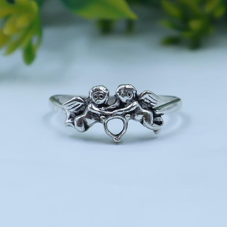 May include: A silver-toned ring featuring two cherub figures facing each other with wings extended. The cherubs are connected at the center by a small, open heart-shaped detail. The metal has a polished finish with darkened recessed areas.