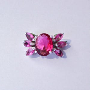 May include: A silver ring featuring a large, oval-cut, magenta gemstone as the centerpiece. The ring is adorned with smaller, petal-shaped, pink gemstones that radiate outwards, creating a floral design. The ring is set against a white background.