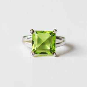 Puede incluir: Un anillo de plata con una gran gema verde claro de talla cuadrada. La gema está sujeta por cuatro garras de plata. El anillo está sobre un fondo blanco.