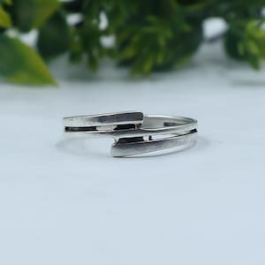May include: A silver-toned ring featuring a bypass design with two parallel bands that overlap at the center. The metal has a smooth, polished finish with darkened recessed areas between the bands, creating a distinct layered appearance.